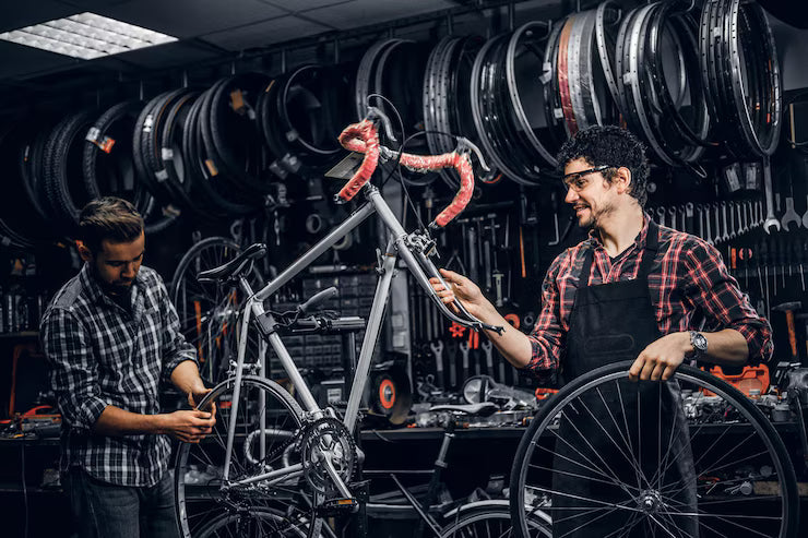Two mechanics working on a bicycle in a workshop with tools and parts visible.
